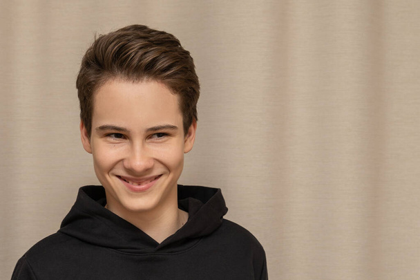 Nice guy 13-14 years old European in black hoodie smiling on a beige background, close-up portrait as a mock-up - Фото, изображение