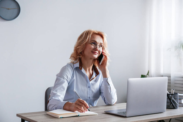 Portrait of adult blonde woman wearing blue blouse sitting at her workplace talking on the phone and smiling. Joyful female having fun phone call in the office. Background, copy space, close up. - 写真・画像