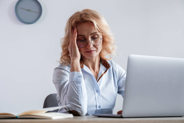 Portrait of adult businesswoman experiencing a massive migraine at her workplace. Anxious woman sitting by the desk at the office touching her head due to a headache. Background, close up, copy space. - Valokuva, kuva