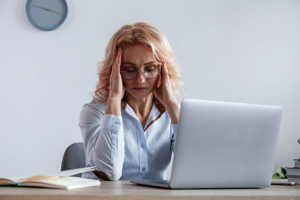 Portrait of adult businesswoman experiencing a massive migraine at her workplace. Anxious woman sitting by the desk at the office touching her head due to a headache. Background, close up, copy space. - Fotografie, Obrázek