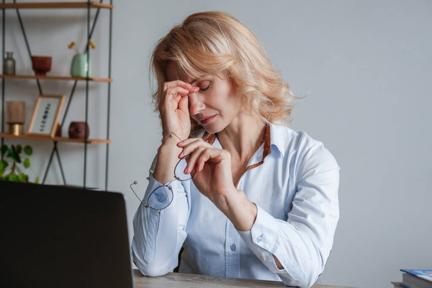 Portrait of adult businesswoman experiencing a massive migraine at her workplace. Anxious woman sitting by the desk at the office touching her head due to a headache. Background, close up, copy space. - Foto, Imagem