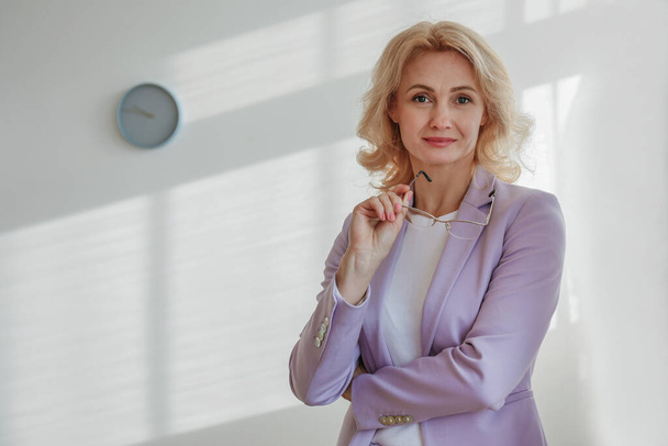 Beautiful blonde businesswoman leaning against the wall with arms crossed over the chest. Confident adult woman wearing lilac jacket standing with folded hands. Office background, copy space, close up - Foto, afbeelding