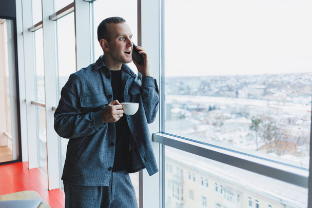 Hezký muž evropského vzhledu v neformálním obleku pije kávu, mluví po telefonu a dívá se z okna - Fotografie, Obrázek