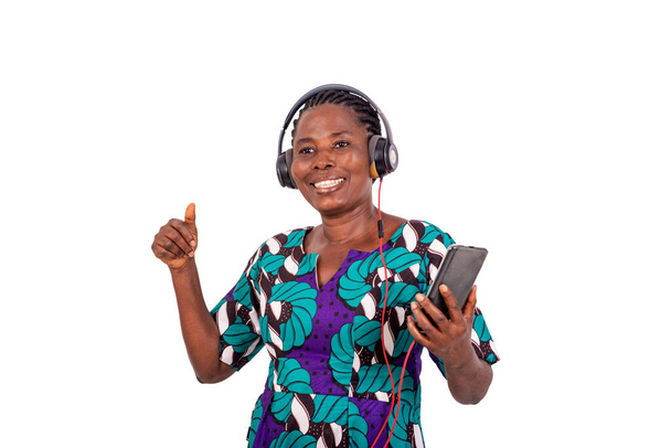 afrikkalainen nainen seisoo valkoisella taustalla kuunnellen musiikkia kännykällä ja kuulokkeilla nauraen. - Valokuva, kuva
