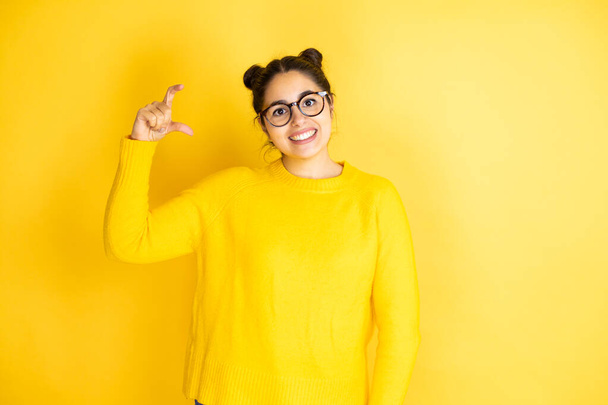 孤立した黄色の背景笑顔の上にカジュアルセーターを身に着けている若い美しい女性と指で小さなサイズの看板を行う手で自信を持ってジェスチャー。概念の測定. - 写真・画像