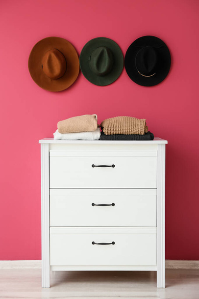 Ζεστά πουλόβερ σε συρταριέρα και κομψά καπέλα που κρέμονται σε ροζ τοίχο - Φωτογραφία, εικόνα