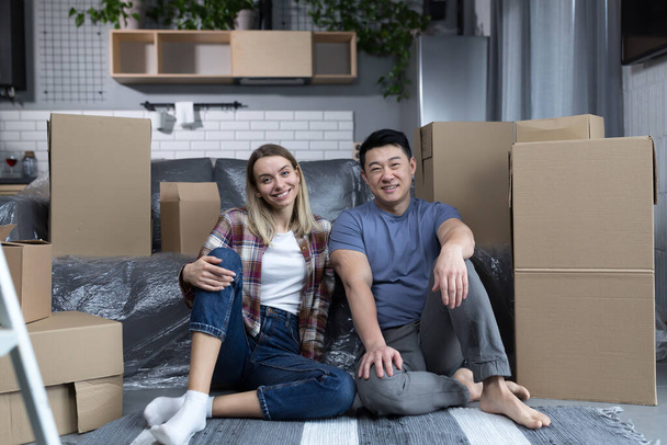 Young couple man and woman hugging together happy looking at camera, sitting on the floor of a new apartment, happy multiracial family - Фото, изображение