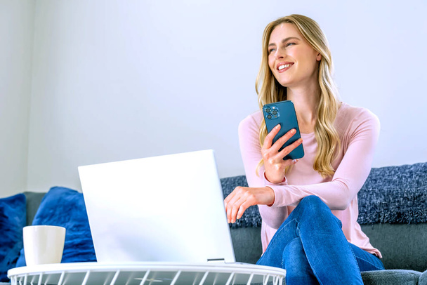 Szép, szőke hajú nő dolgozik a laptopon, miközben otthon ül a kanapén.  - Fotó, kép