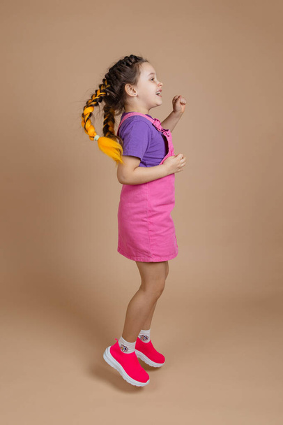 Energetic female kid in process of jumping with two kanekalon pigtails of yellow color wearing pink jumpsuit, purple t-shirt and pink sneakers on beige background - Zdjęcie, obraz