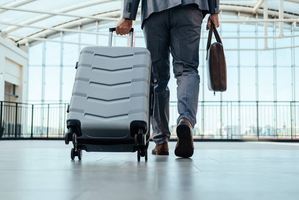 Vista de ángulo bajo del hombre de negocios irreconocible en elegante traje gris que camina a través de la terminal del aeropuerto con maletín y maleta - Foto, Imagen