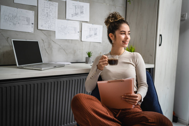 одна жінка молода біла жінка сидить за столом на роботі в офісі навчальні документи, розслабляючі в той час як тримайте кліп-графік з паперами реальні люди копіюють простір жінка-інженер або студент насолоджується кавою
 - Фото, зображення