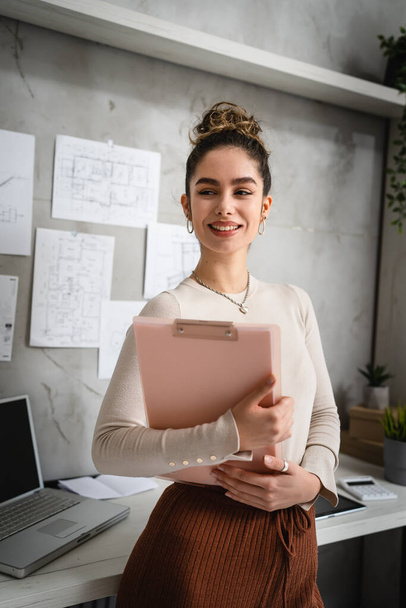Front view portrait of one woman young female student or entrepreneur or manager standing in front of the desk at her office or home holding documents looking to the side smile happy real people - Photo, Image