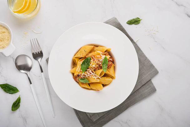 Bolognese Penne Pasta. Tasty appetizing classic italian penne pasta with parmesan cheese, basil and Bolognese sauce in white plate on plate on stone table. Traditional dish of Italian cuisine Top view - Zdjęcie, obraz