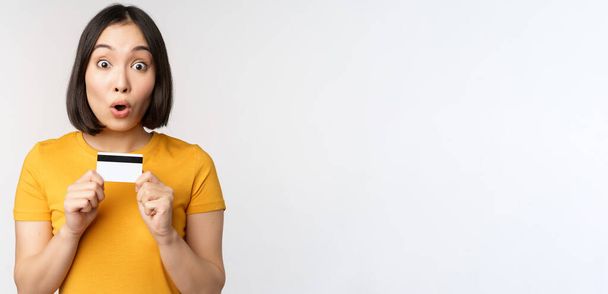 Retrato de bela menina coreana segurando cartão de crédito, recomendando serviço bancário, de pé em camiseta amarela sobre fundo branco - Foto, Imagem