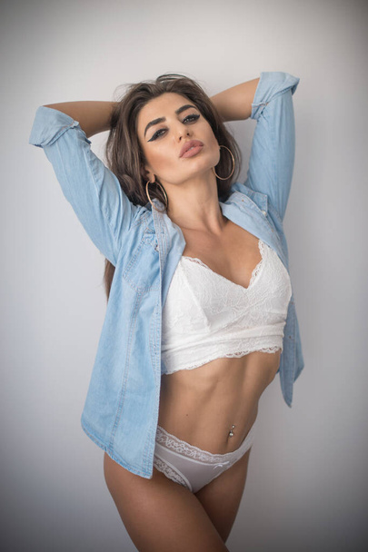 Attractive sexy brunette in white lingerie and denim shirt posing provocatively, near a grey wall,studio shot. Portrait of sensual woman with long hair, in classic boudoir scene. - Foto, Bild
