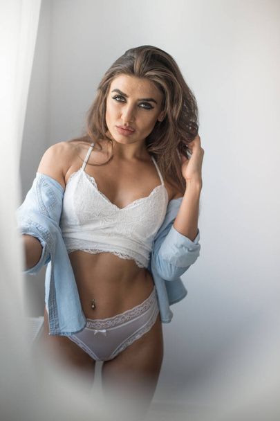 Attractive sexy brunette in white lingerie and denim shirt posing provocatively, near a grey wall,studio shot. Portrait of sensual woman with long hair, in classic boudoir scene. - Foto, imagen