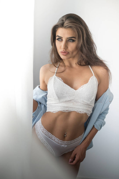 Attractive sexy brunette in white lingerie and denim shirt posing provocatively, near a grey wall,studio shot. Portrait of sensual woman with long hair, in classic boudoir scene. - Photo, image