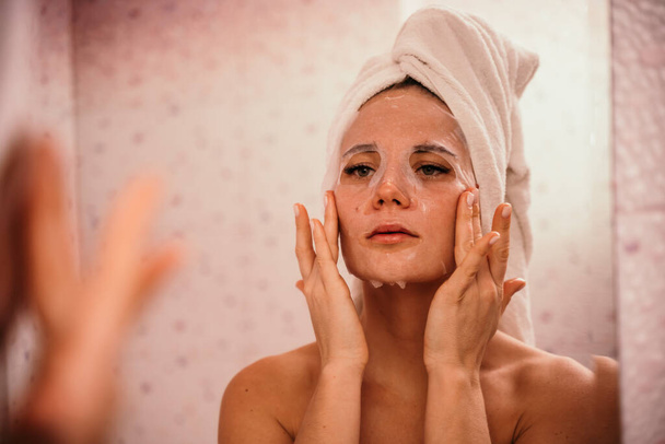 Young beautiful woman using a moisturizing facial mask after taking a bath. Pretty attractive girl in a towel on her head stands in front of a mirror in a home bathroom. Daily hygiene and skin care - Photo, Image