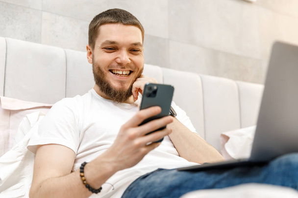 молодой человек дома на диване с ноутбуком и телефоном - Фото, изображение