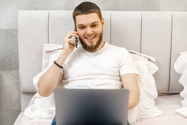 Nuori mies kotona sohvalla puhumassa puhelimessa ja katsomassa kannettavaa tietokonetta - Valokuva, kuva