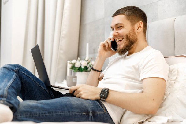 Молодой человек дома на диване разговаривает по телефону и смотрит на ноутбук - Фото, изображение