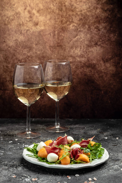 mediterranean salad with prosciutto or jamon, mozzarella and green basil leaves and Cantaloupe melon on white table, traditional Spanish and Italian appetizer served with wholemeal grissini, top view. - Foto, Imagem