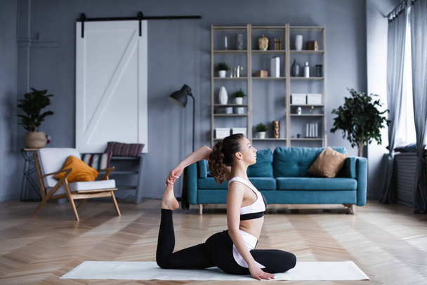Fiatal, sportos nő otthon edz, nyújtózkodik, miközben egy jóga szőnyegen ül a nappaliban. Otthoni edzés. - Fotó, kép