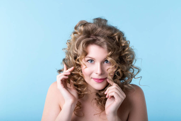 Young beautiful woman with curly hair smiles on a blue background. Female emotional portrait in the studio. - Valokuva, kuva