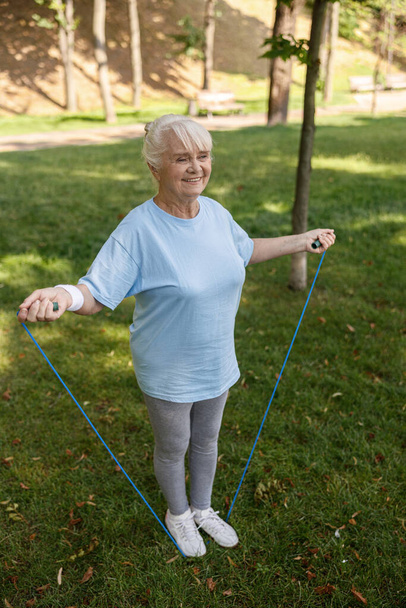 Spor giyim bölümündeki mutlu yaşlı kadın yeşil parkta ip atlama egzersizi yapıyor. - Fotoğraf, Görsel
