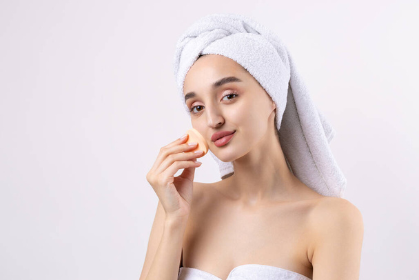 Beautiful girl with thick eyebrows and perfect skin at white background, towel on head, beauty photo. Holds a cosmetic sponge. - Фото, зображення