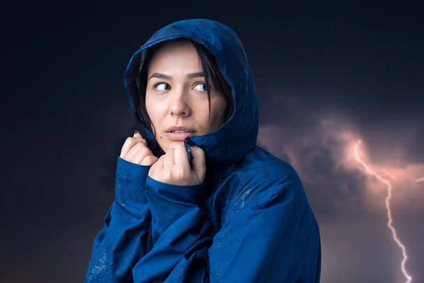 Πορτρέτο ενός χαμογελαστού κοριτσιού ντυμένο με μπλε αδιάβροχο σε σταγόνες ποζάρουν με κουκούλα στο φόντο ενός ζοφερού ουρανού με αστραπή. - Φωτογραφία, εικόνα