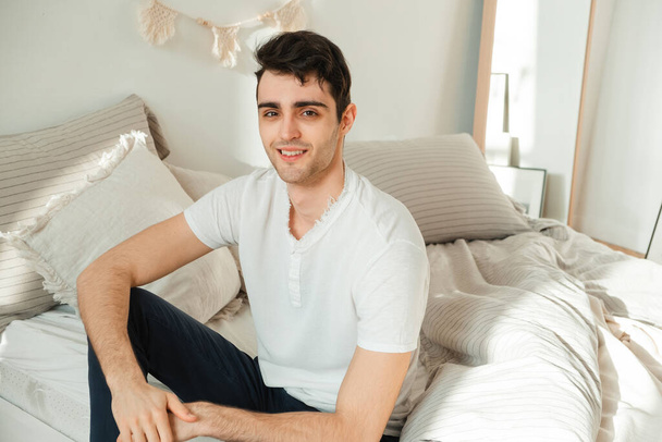 Muotokuva iloinen nuori mies rentoutuu makuuhuoneessaan, istuu sängyllä ja katselee kameraa - Valokuva, kuva