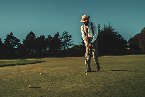 View of a fancy mature African guy in an elegant retro outfit with a straw hat and a tie hitting the ball with a club while standing on the lawn of the private golf club on a warm sunny evening - Photo, image