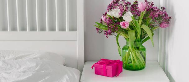 Pink box present and vase with spring flowers on bedtable near bed. Scandinavian Interior. Banner image for web page, design - Photo, Image