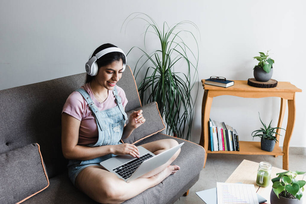 メキシコの自宅でソファに座っている間、ビデオ通話やオンラインクラスでラップトップを使用してヘッドフォンを持つ若いラテン女性の学生  - 写真・画像