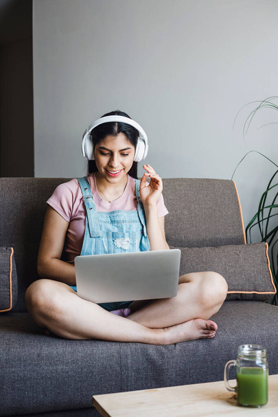 joven estudiante latina con auriculares usando portátil en una videollamada o clase en línea mientras está sentada en un sofá en su casa en México  - Foto, imagen