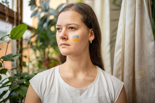 Ein trauriges junges ukrainisches Mädchen mit der Fahne der Ukraine auf dem Gesicht blickt durch das Fenster. Das Konzept der Beteiligung des ukrainischen Volkes am Krieg gegen Russland. Kein Kriegskonzept - Foto, Bild
