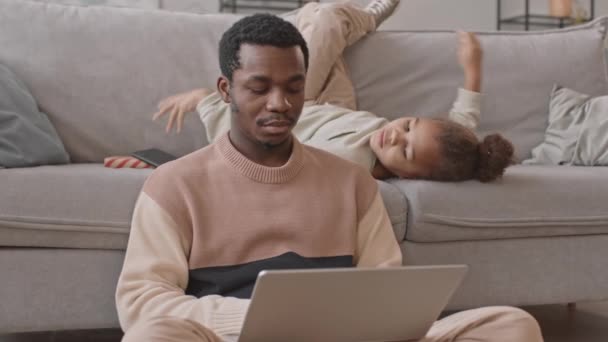 中出しの忙しいアフリカ系アメリカ人男性タイプのラップトップで自宅から仕事中にロックダウンと彼の8歳の遊び心のある娘から仕事から彼をそらす - 映像、動画