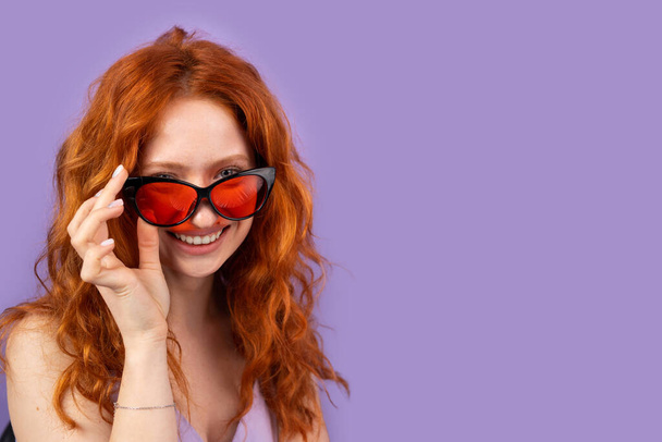 Young redhead woman in spring summer dress smling widely taking off her red sunglasses on purple background and copy space for advertising. - Foto, Imagen