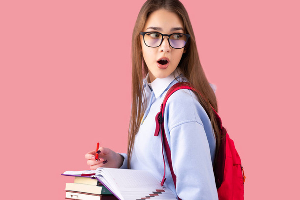 Amazing school girl in uniform holding a stack of books and a pen looking over her shoulder with open-eyed expression. - Фото, изображение