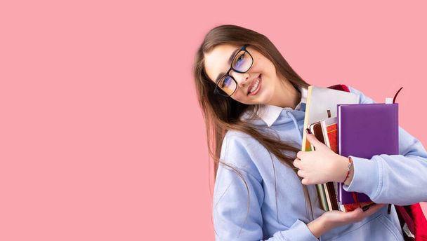 Lustig lächelndes Schulmädchen mit Rucksack und Büchern in der Hand in Uniform mit Brille, die glücklich auf rosa Hintergrund und Kopierraum schaut. - Foto, Bild