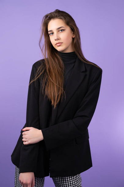 Serious pretty blond young woman with make-up wearing black coat and checkered trousers on purple background, people and education concept. - Foto, afbeelding