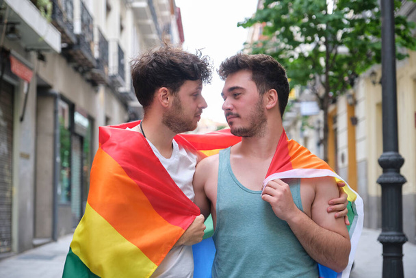 Gay couple wrapped in a LGBT flag, hugging and looking each other. - Photo, Image