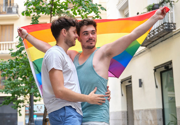 Гей-пара улыбается и обнимается с флагом ЛГБТ. - Фото, изображение