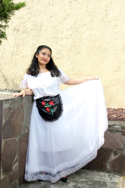 Молодая девушка-подросток из Веракруса носит белое платье с черным вышитым фартуком, готовым танцевать, гордясь своей культурой и традициями, с каменной стеной и деревом - Фото, изображение