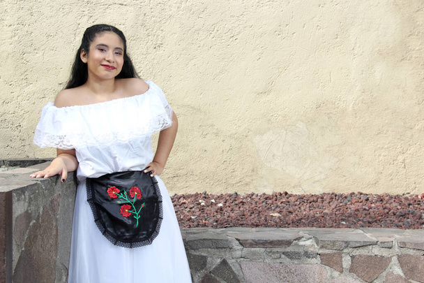 Junge Teenagerin aus Veracruz trägt ein weißes Kleid mit schwarzer bestickter Schürze, die bereit ist, stolz auf ihre Kultur und Tradition mit einer Steinmauer und einem Baum zu tanzen - Foto, Bild