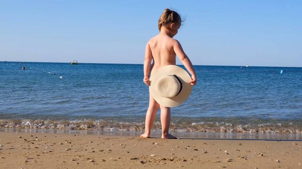 zabawna dziewczynka 3 lata w słomkowym kapeluszu patrzy na morze, widok z tyłu, zbliżenie. Dzieciństwo, związek, wakacyjny pomysł. selektywna koncentracja na kapeluszu - Zdjęcie, obraz