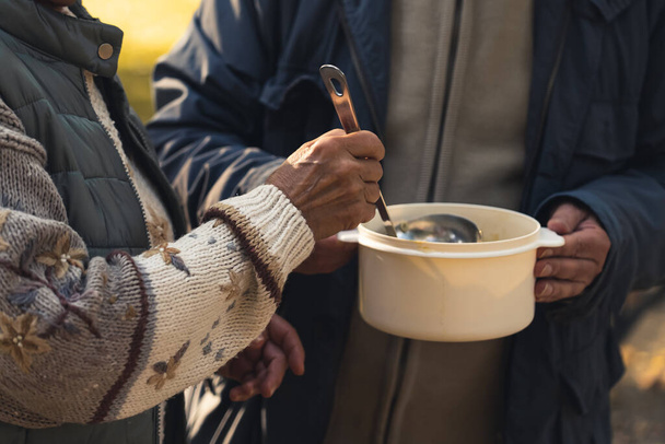 Sharing warm food with people in need concept. Cooking pot full of soup is handled between homeless people who share it with each other. - Photo, Image
