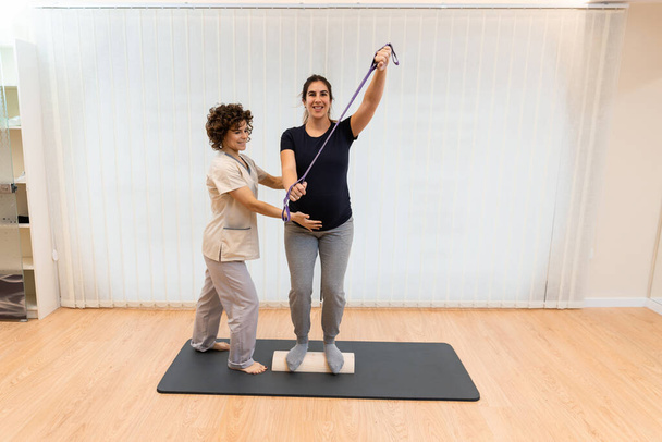 Těhotná žena stojící na proprioceptivním kufru nebo dřevěném válečku s elastickým proužkem s fyzioterapeutem - Fotografie, Obrázek