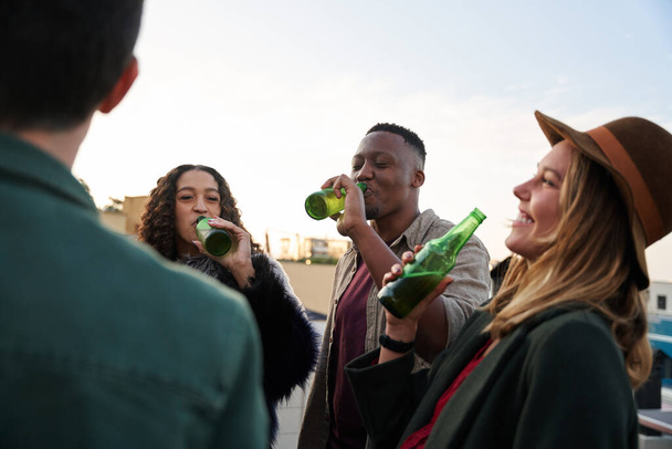 Multi-cultural group of young adult laughing with drinks on the balcony of a rooftop terrace in the city.  - Fotó, kép